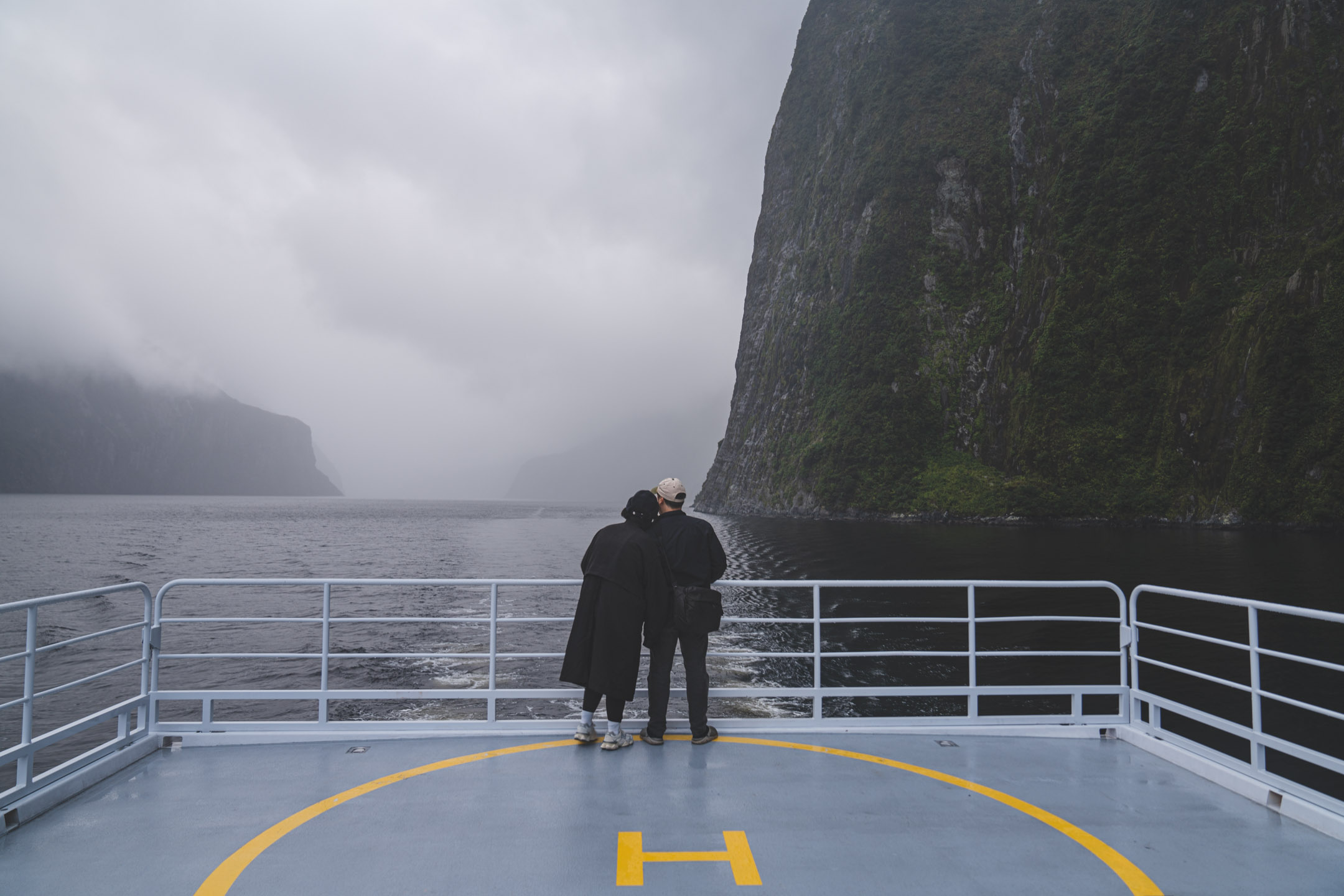 Two travellers at the stern rail of a ferry in Milford Sound, cliffs vanishing into cloud